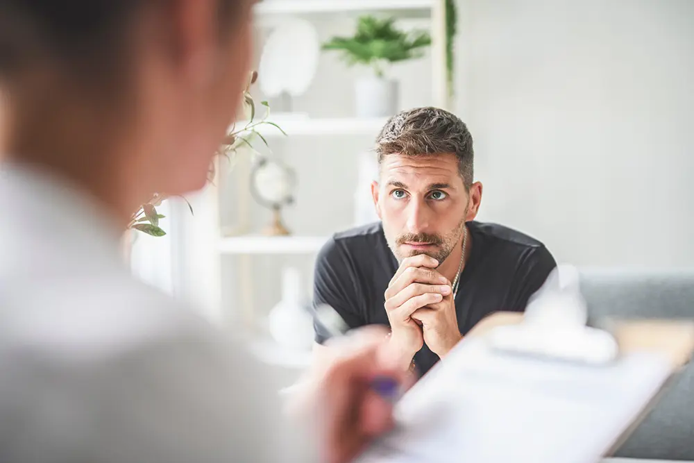 A man with light brown hair and a beard sits with his hands clasped, looking intently at a person holding a clipboard in the foreground, suggesting a serious conversation or counseling session in a bright room.
