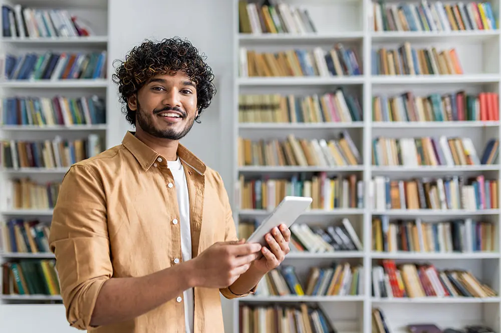 A smiling person with curly hair and a brown shirt holds a tablet while standing in front of bookshelves filled with books in a library or study room.