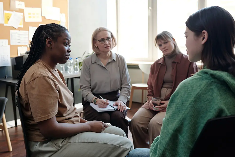 Young African American woman, mature female psychologist and blond girl looking at brunette patient in green pullover in counseling session