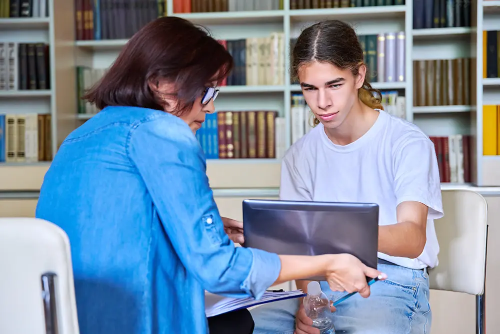 A woman and a teenage boy sit together in a library, looking at a laptop screen. The woman points at something on the laptop while the boy listens attentively. Bookshelves are visible in the background.