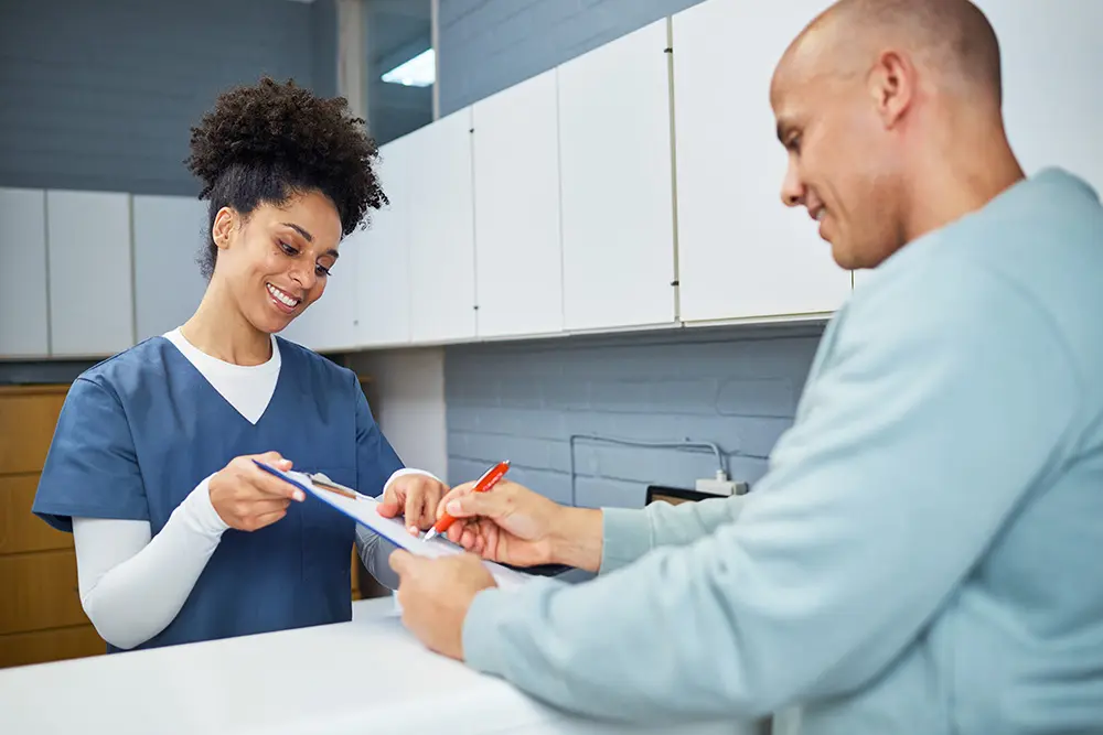 A smiling healthcare worker in scrubs hands a clipboard to a man at a reception desk, while the man signs a form with a red pen. White cabinets are visible in the background.