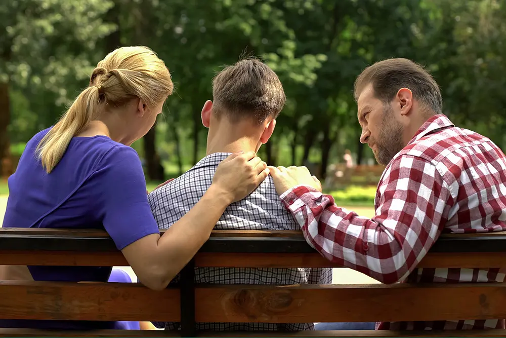 Three people sit on a park bench, two adults comforting a younger person sitting between them by placing hands on his back and shoulder. The scene is outdoors with trees in the background.