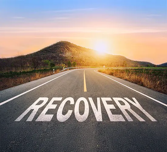 A paved road leads toward a hill at sunrise, with the word RECOVERY painted in large white letters on the asphalt, symbolizing a journey or path to recovery.